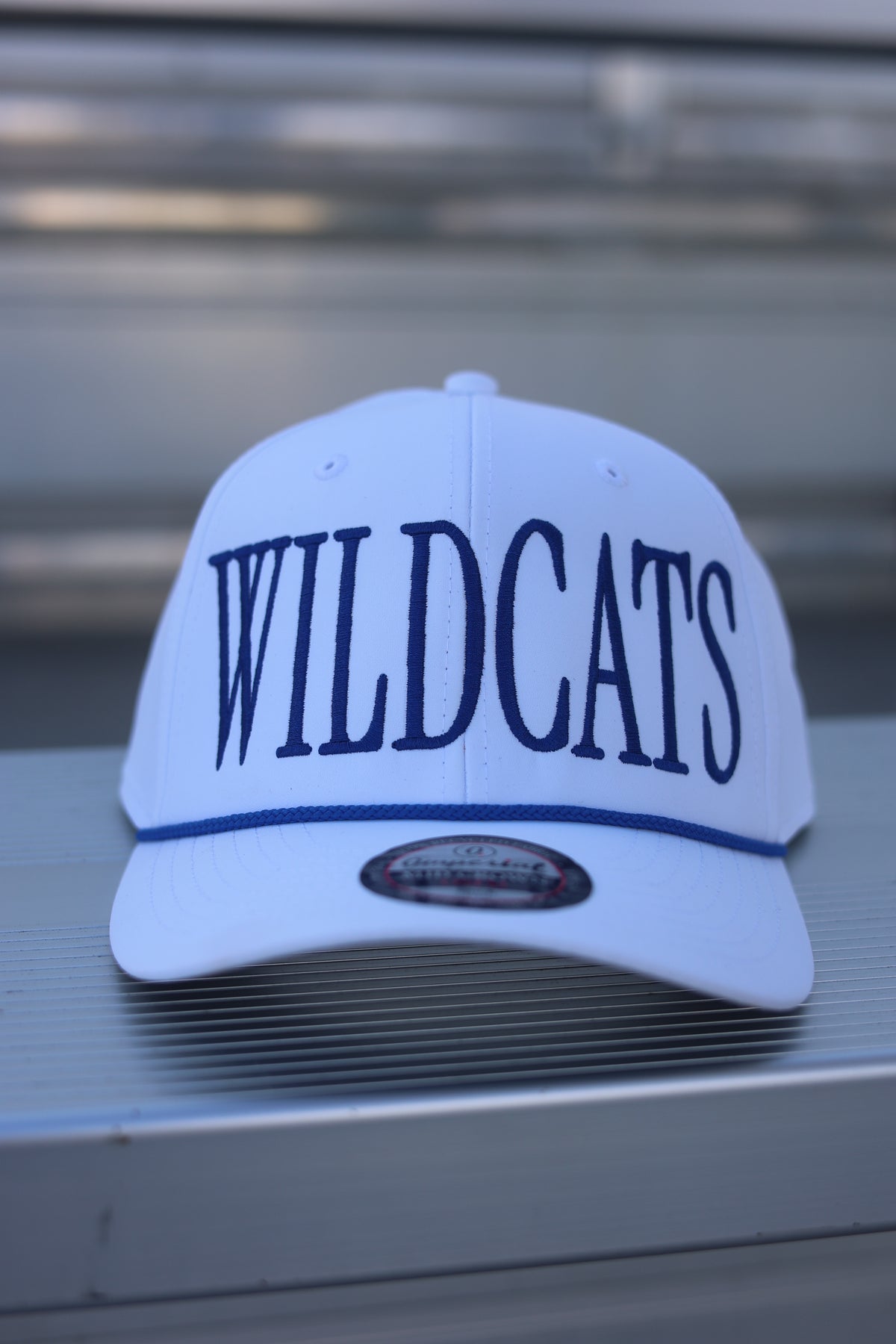 A white baseball cap with 'WILDCATS' embroidered in navy blue across the front, resting on a gray metal surface.